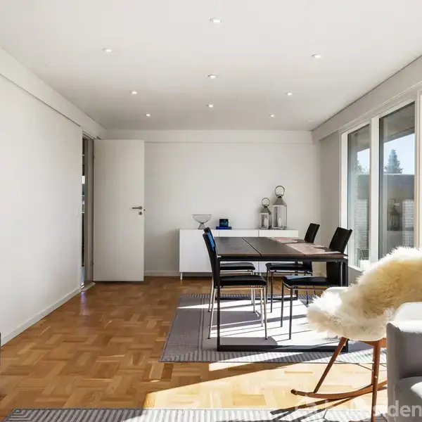 Spisestue med et moderne bord og sorte stole. Rummet har trægulv og store vinduer til højre, der lukker lys ind. En hvid dør er i baggrunden, sammen med minimalistisk indretning.