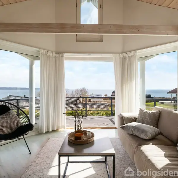 Stue med sofa og stol, omgivet af store vinduer med udsigt over havet. En kaffe i midten og bløde puder skaber en hyggelig atmosfære.
