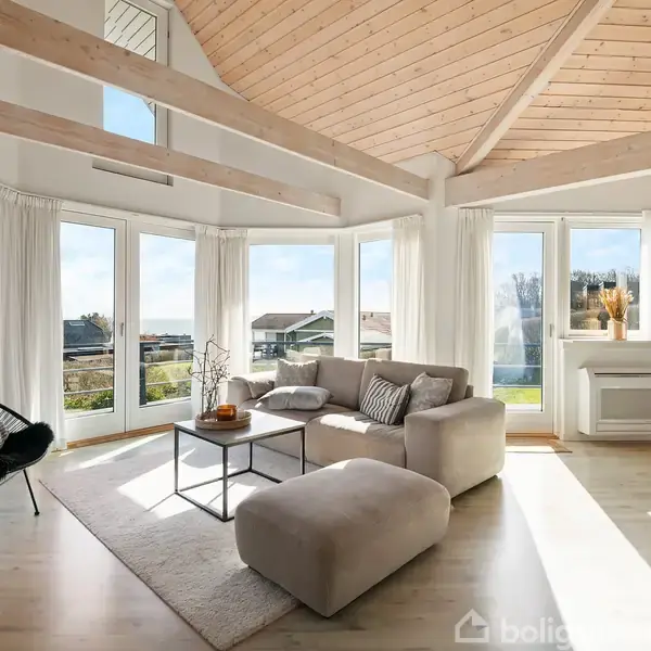 Et lyst, moderne stueindretning med træloft og store vinduer. En beige sofa og skammel står på et lyst tæppe. Udenfor ses et grønt landskab og blå himmel.