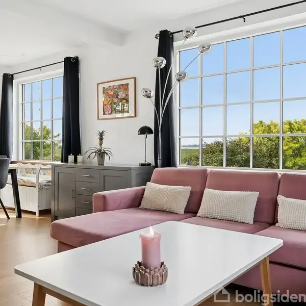 En lyserød sofa står foran et stort vindue med udsigt til træer. Et hvidt sofabord med pynt er foran sofaen. Lys, moderne stue med spisebord i baggrunden.