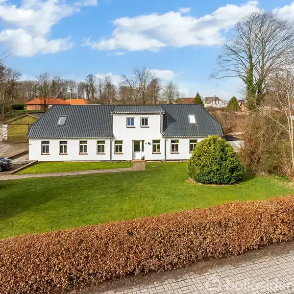 Hvidmalet hus med skråt tag, placeret i et rummeligt, velplejet haveområde med græsplæne og buske. Omgivet af træer og mindre bygninger i et boligområde.