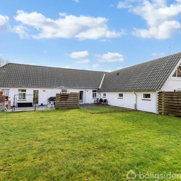 En stor hvid villa med skråtag præsenterer en velholdt have med grøn græsplæne. Baggrunden viser en klar blå himmel med spredte skyer.