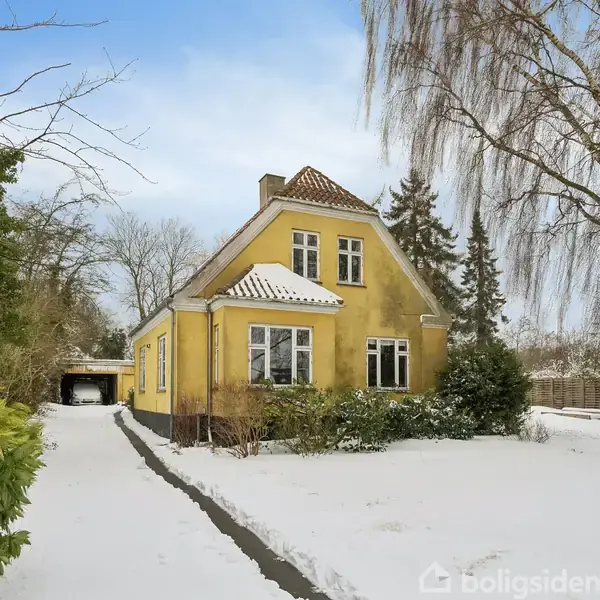 Gulmalet hus står stille i en snedækket have med en sti, der fører til en garage. Omgivet af træer og buske under en lys, skyet himmel.