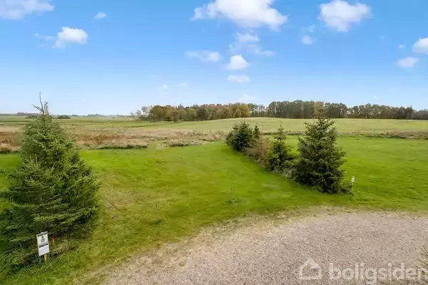 Parceller omgivet af grønne græsplæner og flere træer, placeret i et landligt område med en klar blå himmel og spredte skyer. En grusplads er synlig i forgrunden.