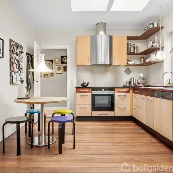 Et moderne køkken har lyse træskabe, en ovn med emhætte, og er omkranset af en spiseplads med et rundt bord og farverige skamler. Væggene har billeder og opslagstavle.