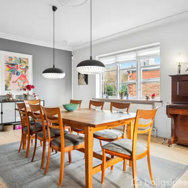 Et spisebord med otte stole står på et tæppe i en spisestue. To lamper hænger over bordet. Til højre står et klaver, og til venstre ses et skab og et maleri. En stor vindue giver udsigt til en murstensbygning.