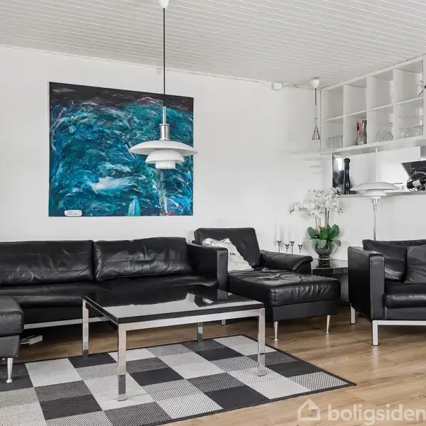 Sort lædersofa sæt med sofabord står på et mønstret tæppe i en moderne stue. Kunstmaleri hænger på væggen; køkken ses i baggrunden.