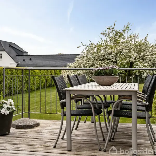 Et træbord med sorte stole står på en terrasse. En blomstrende busk og grøn plæne omkranser terrassen i et boligområde. En potteplante står ved siden af bordet.