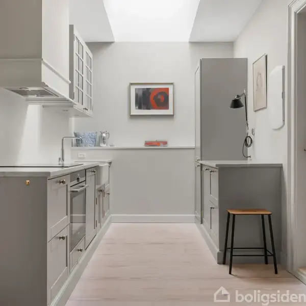 Et moderne køkken med grå skabe står tomt. Et lille bord med en stol ved siden, belysning fra ovenlysvindue. Væggen er dekoreret med et billede og lamper.