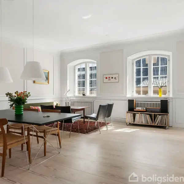 Stue med spisebord og stole i forgrunden, blomster i vase. Sofaområde ved siden af vinduer med udsigt. Hylde fyldt med plader nær radiatoren og væglamper. Lumineus interiør.