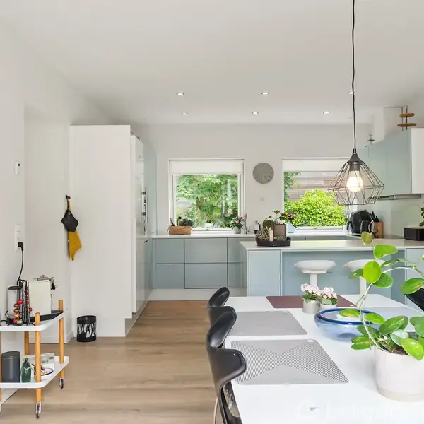 Køkkenbord med spiseplads, omgivet af moderne køkkenskabe i et lyst rum. Under hænger en geometrisk lampe, mens planter og pynt dekorerer bordet. Vinduer viser udsigt til have.