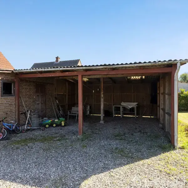 Carport med grusbelægning; indeholder tre cykler og haveudstyr. Omgivet af en murstensbygning med rødt tag og græsplæne i baggrunden. Solrig dag med klar blå himmel.