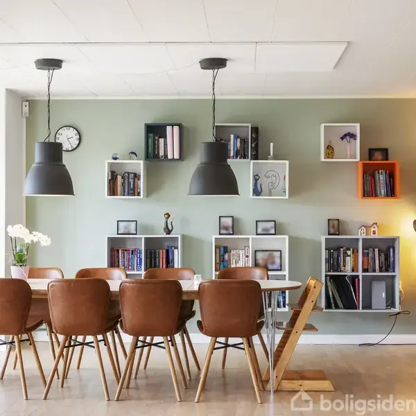 En stue med en brun hjørnesofa står ved et vindue. Bordet foran sofaen har bøger. Rummet er lyst og moderne, set gennem åbne dobbelte franske døre.