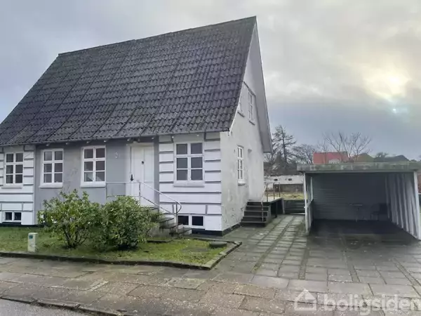 Et hvidt to-etagers hus med skiferet tag står på en gade ved siden af en åben garage. Foran huset er en lille græsplæne og buske. Himlen er overskyet.