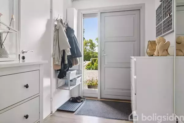 En lys entré med en hvid dør til haven. Jakker hænger på knager til venstre, og der står sko på en måtte ved døren. Pejsefigurer på en skuffe.