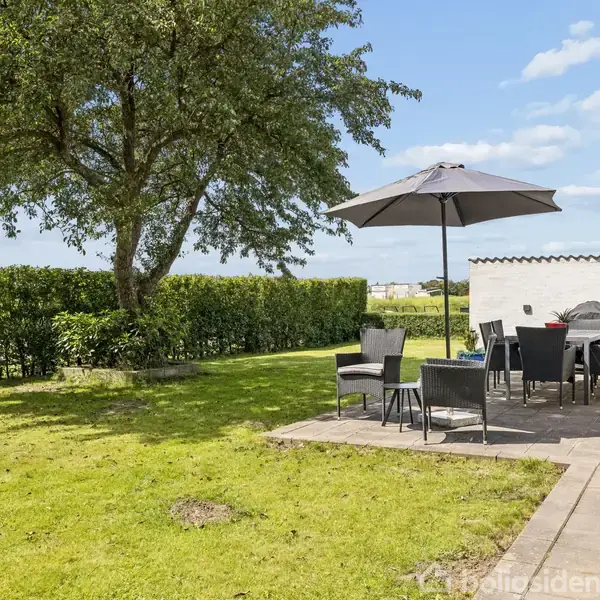 Haveområde med terrasse, hvor sorte havemøbler og en parasol står under et stort træ. En hæk løber langs kanten af græsplænen.