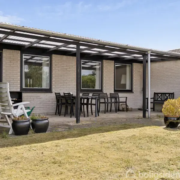 Huset har en overdækket terrasse med spisebord og stole, omgivet af en velplejet græsplæne og høje hække. Flisebelægning og krukker med planter skaber en hyggelig udendørs atmosfære.
