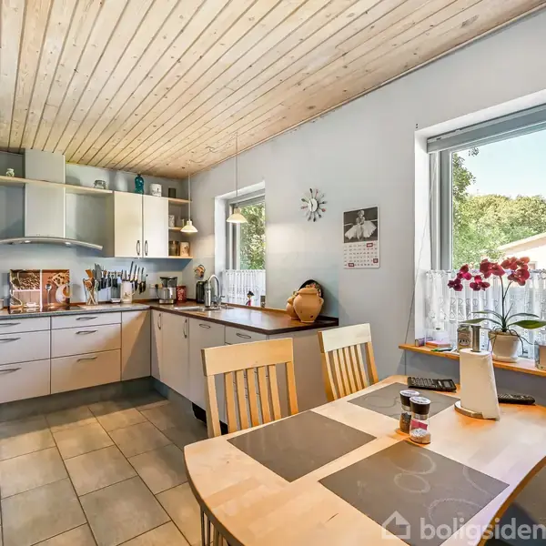 Et moderne køkken med lys træloft og grå fliser, hvor spisebordet står ved vinduet med udsigt til have. Køkkenet har hvide skabe, komfur og diverse køkkenudstyr.