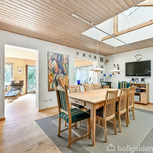 Spisebord omgivet af træstole står på et tæppe i en spisestue. Over bordet hænger lamper, og der er kunst og et tv på væggene. Gulv og loft er af træ.