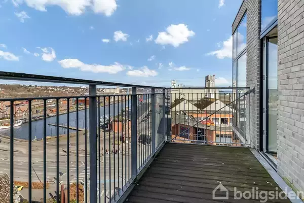 En altan med træbund og metalrækværk vender mod en by, hvor man kan se en flod, bygninger og en klar blå himmel.