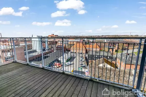 Altan med metalrækværk vender mod en byudsigt med rødstensbygninger og fabrikker under en blå himmel med spredte skyer. Ældre byområde i baggrunden.