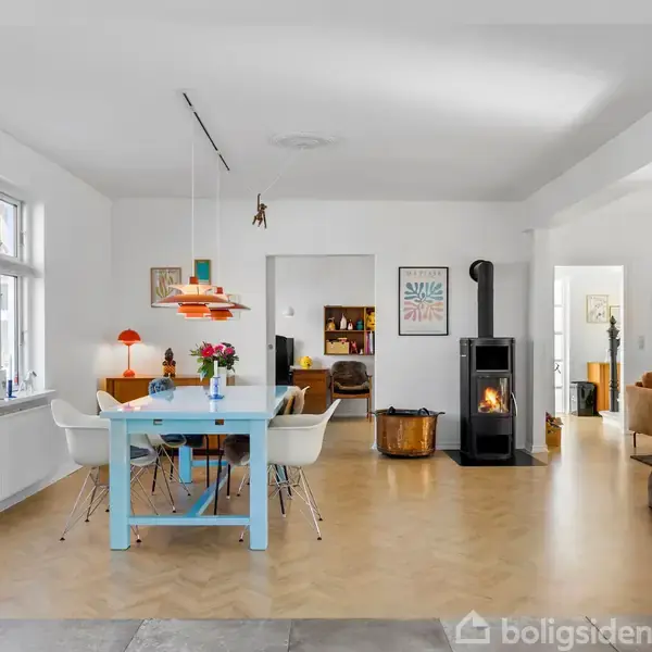 Et moderne køkken-alrum med et lyseblåt spisebord og hvide stole, omgivet af hvidmalede vægge og store vinduer. En brændeovn står på trægulvet i baggrunden.