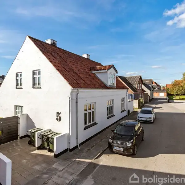 Hvidt hus med rødt tegltag og nummer 5 på facaden står stille, flankeret af to parkerede biler på villavej. Solrigt vejr og blå himmel danner baggrund.