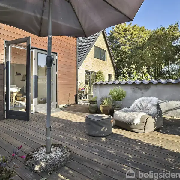 En træterrasse med parasol og åbne terrassedøre leder ind til et hus. Man ser en blød stol med et tæppe og planter i krukker bagved i solrig haveomgivelse.
