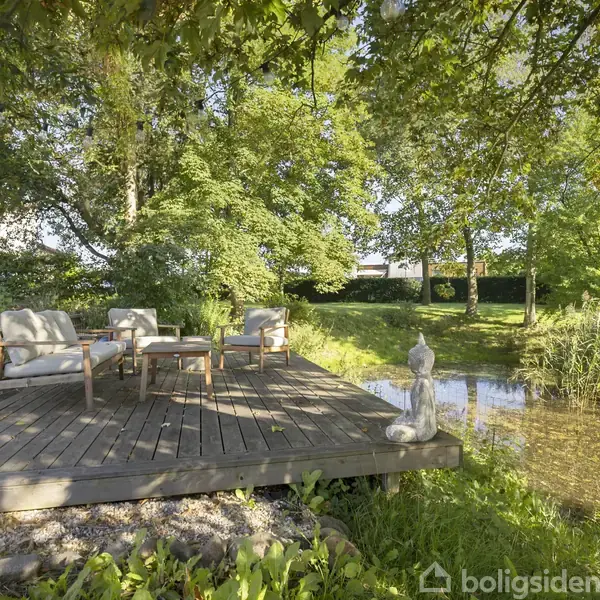 Trædæk med to polstrede stole og et lille bord står ved en rolig dam, omkranset af frodige grønne træer. En statue ses i nærheden af vandkanten.