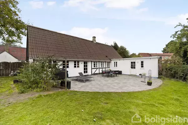 Hus med rødbrunt tag står i en have med græsplæne. Terrasse foran huset har liggestole og bord med stole. Sollys belyser området, der er omgivet af træer og buske.