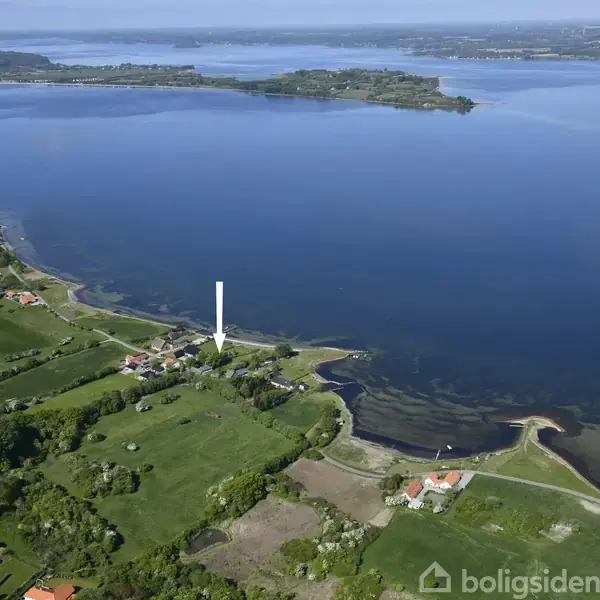 En kystnær landsby ses fra luften med spredte huse og frodige marker i forgrunden. En stor, blå fjord strækker sig i baggrunden, med skovklædte områder omkring.