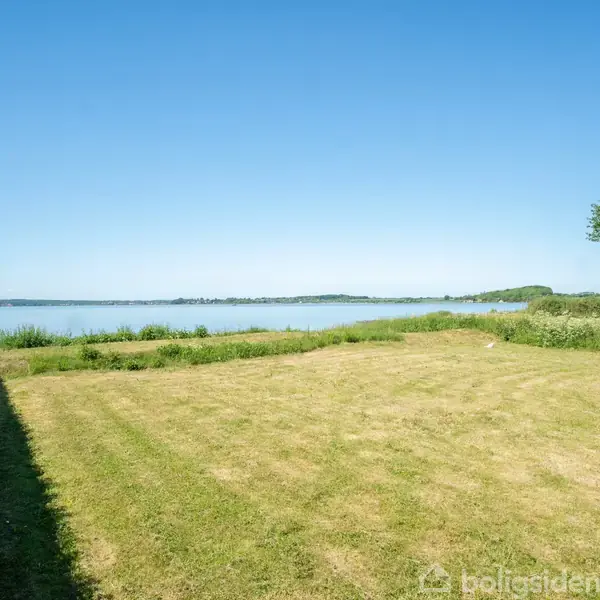 Græsplæne strækker sig mod en sø, der omgives af grøn bevoksning under en klar blå himmel. Hæk til venstre og træer til højre rammer scenen ind.