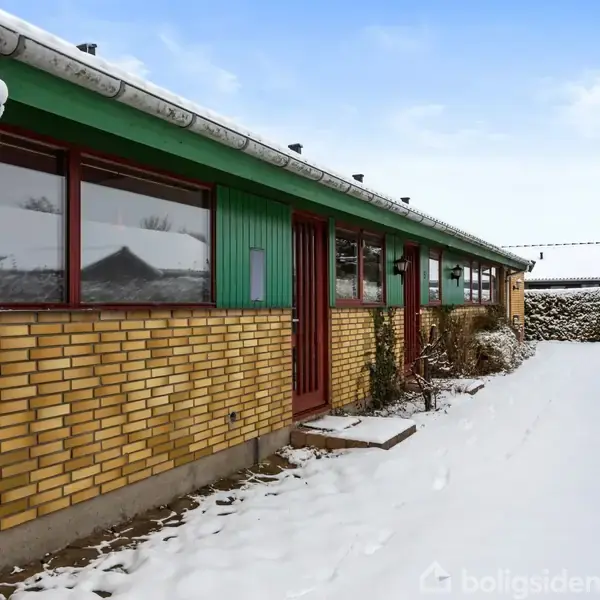 En gul murstensbygning med røde døre og vinduer står i sne fyldt forhave. Hækken i baggrunden danner en grøn ramme under den blå himmel.