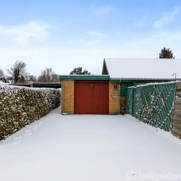 Garage med rød port står i en gårdsplads dækket af sne. Hæk til venstre og hegn til højre omgiver indkørslen. Himlen er blå og klar.