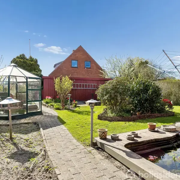 Drivhus står ved siden af en brolagt sti i en velplejet have med en lille dam. En rød bygning med trekantet tag er i baggrunden under en blå himmel.