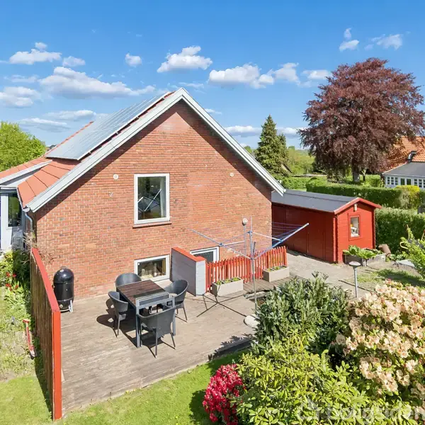 Rødt murstenshus med sort tag, omgivet af have. Foran huset er en terrasse med havemøbler og grill. Solskinsdag med få skyer på himlen og grønne buske omkring.