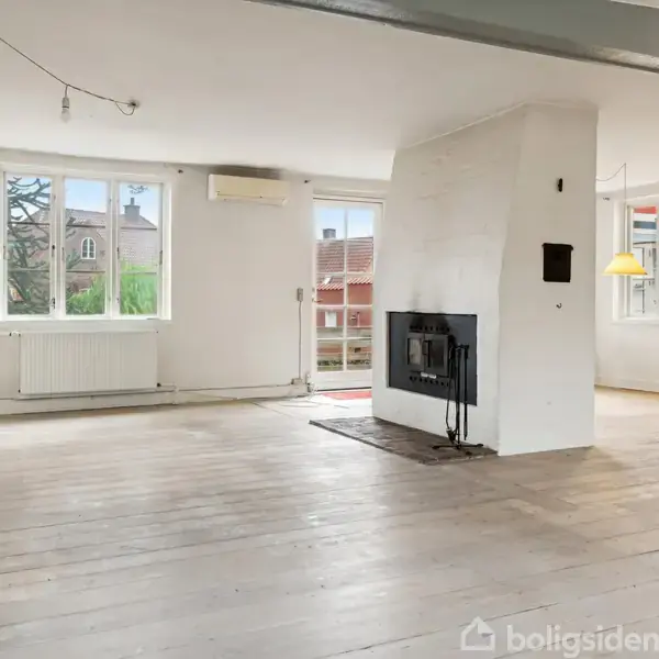 En rummelig stue med trægulv, hvor pejsen står centralt. To store vinduer slipper lys ind fra udsigten over hustage. Forskellige lamper hænger fra loftet. Ingen tekst til stede.