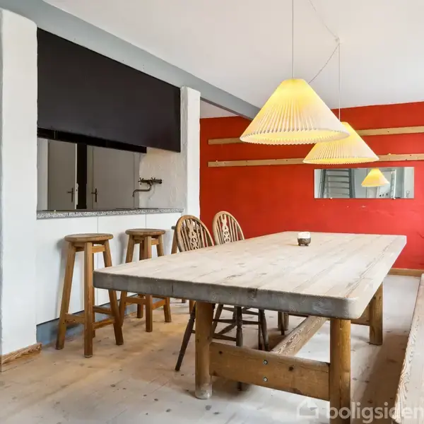 Langt spisebord med træbænk og stole står i et rum med en rød væg. To gule lamper hænger over bordet, og der er et lille serveringsvindue.