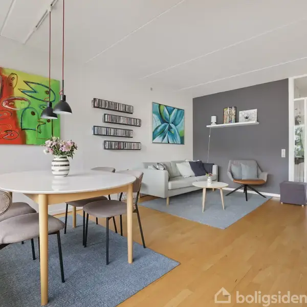 Et spisebord med fem stole står på et tæppe i en moderne stue. Kunst hænger på væggen, og en sofa med bord er i baggrunden. Blomstrende vase på bordet.