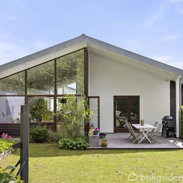 Et moderne hus med glaspartier og træterrasse står i en velplejet have. Havemøbler er placeret på terrassen, omgivet af planter. Himlen er blå med få skyer.