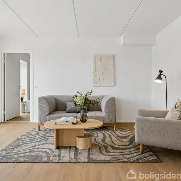 En stue med to grå sofaer står omkring et rundt træbord med en vase og bøger. En gulvlampe til højre; vægkunst hænger bagved. Store vinduer til venstre.