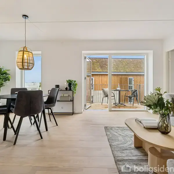 Spisestue med bord og fire stole, hængende lampe. Sofa og sofabord med planter på tæppe. Store vinduer åbner til en terrasse med to stole og bord.