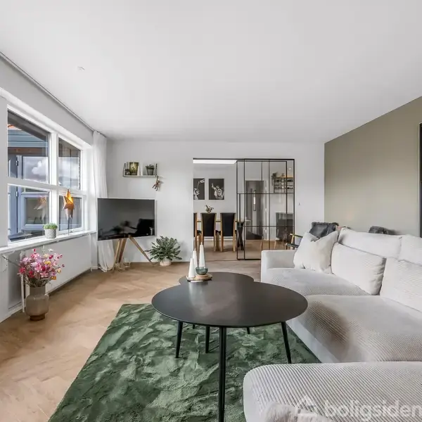 En lys stue med en stor grå sofa står ved siden af et sort rundt sofabord på et grønt tæppe. Store vinduer til venstre giver udsigt til en terrasse.