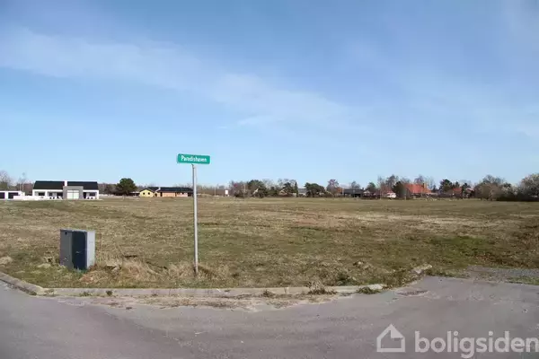 Et vejskilt med teksten "Pindsvineø" står ved en tom græsmark omgivet af spredte huse og træer under en blå himmel.