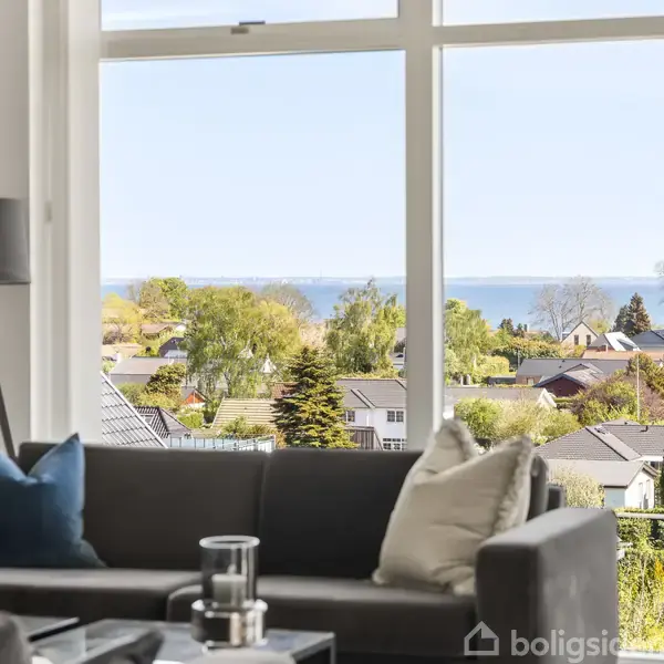 Moderne hvidt hus med store vinduer, beliggende på en frodig grøn græsplæne omgivet af buske og træer. En terrasse med havemøbler er ved siden af huset.