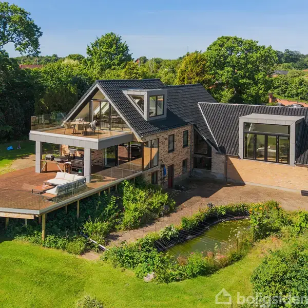 En moderne loftstue med skråvægge og synlige træbjælker har en stor vinduesfacade, der åbner op til en terrasse. Rummet er møbleret med en stor sofa, to lænestole, et bord og en skærm.