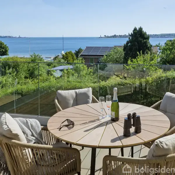 Hus med solpaneler står stille i forgrunden. Udsigt over en rolig bugt og horisonten i baggrunden. Spredt vegetation og nabohuse ses omkring.