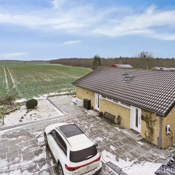 Et gult murstenshus står ved en sneklædt indkørsel, hvor en hvid bil er parkeret. Baggrunden viser åbne, snedækkede marker under en klar, blå himmel.