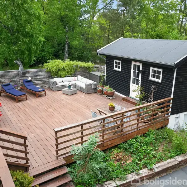 Et træhus står ved en stor terrasse med liggestole og planter. Terrassen er omgivet af grønne træer og buske, hvilket skaber en rolig haveatmosfære.