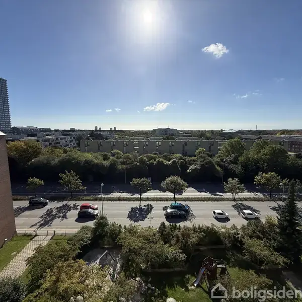 Vejen strækker sig gennem billedet med biler i bevægelse, omgivet af træer og bygninger under en klar blå himmel med solskin.